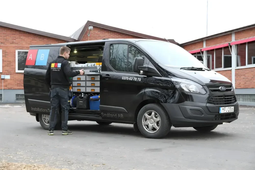 En elektriker organiserar verktyg på baksidan av en svart arbetsbil med öppen sidodörr, parkerad nära en röd tegelbyggnad.