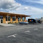 En träbyggnad med en "Jureskogs"-skylt står fridfullt, omgiven av parkerade bilar på en lugn tomt. En elfirmas skåpbil är subtilt synlig under den delvis molniga himlen och antyder elektriker som är upptagna på jobbet i närheten.