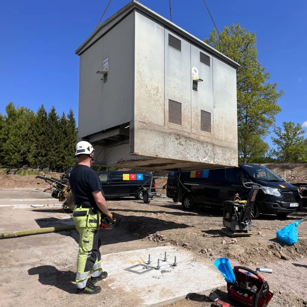 Elektrikern övervakar på ett ansvarsfullt sätt installationen av en stor verktygslåda upphängd i en kran, med skåpbilar och utrustning från elfirman strategiskt placerad i närheten.